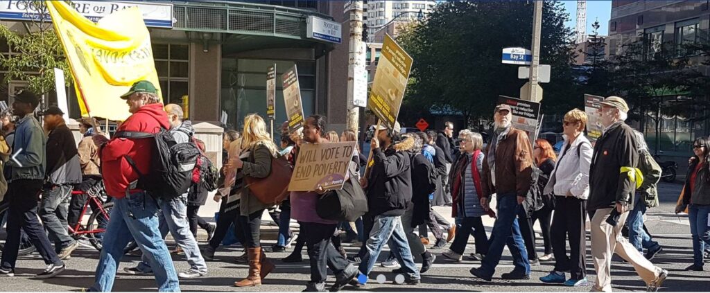 Side view of people walking in a group to the left. Some are holding signs and posters with slogans on them.
